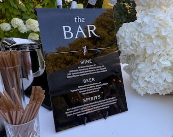 May include: A black sign with the text "the BAR" and a list of wine, beer, and spirits. The bar is set up outdoors with bottles of wine and a floral arrangement. A glass of straws is on the counter.