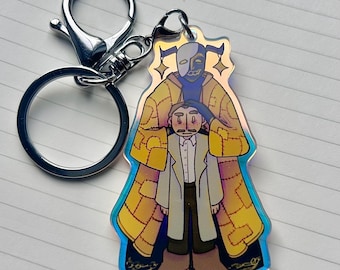 May include: A colorful iridescent keychain featuring a cartoon illustration of two figures. The larger figure is wearing a yellow and blue patterned robe and a mask with a white and black design. The smaller figure is wearing a white shirt, a brown coat, brown pants, and brown shoes. The keychain has a silver metal ring.
