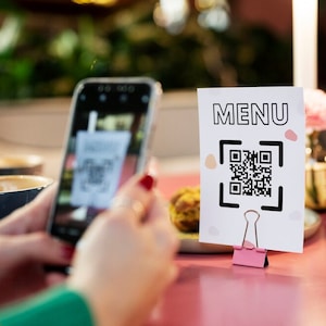 Puede incluir: Un cartel blanco con la palabra "MENU" en texto negro y un código QR en blanco y negro. El cartel está sujeto por un clip de carpeta rosa sobre una mesa rosa. Una persona está usando un teléfono inteligente para escanear el código QR.