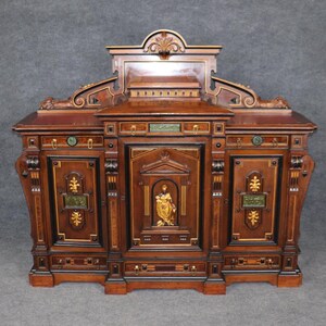 May include: A large, intricately carved wooden cabinet with three doors and a decorative top. The cabinet is made of dark wood with gold accents and inlaid designs. The doors are each decorated with a different design, including a central panel with a figure in relief.