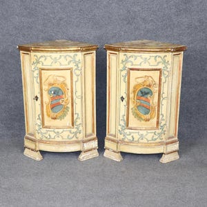 May include: Two antique corner cabinets. Each cabinet has a cream-colored finish with gold trim and decorative painted panels. The panels feature a crest design in blue, red, and gold. The cabinets have a triangular shape.