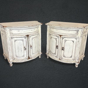 May include: Two white painted antique cabinets with curved fronts and ornate details. Each cabinet has two doors and a single drawer.