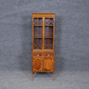May include: A tall, wooden display cabinet with glass doors and a decorative lattice design. The cabinet has two doors on the bottom with oval-shaped wood grain patterns. The cabinet is made of a warm-toned wood and stands on slender legs.