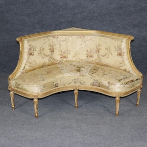 May include: A curved, antique sofa with a gold-painted wooden frame. The sofa features a floral patterned fabric in cream and beige tones. The legs are carved and the back is slightly arched, creating an elegant, vintage aesthetic.