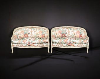 May include: Two cream-colored loveseats with floral patterned upholstery. The sofas have curved backs and armrests, with a white painted wooden frame. The floral pattern includes pink, yellow, and green flowers.