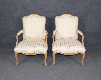 May include: Two antique armchairs with light wood frames and cream-colored plaid upholstery. The chairs have curved arms and legs, and the backs are slightly arched. The overall style is classic and elegant.