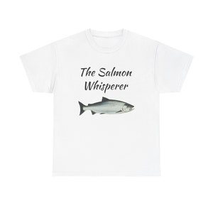 May include: White t-shirt with the text "The Salmon Whisperer" and a black and white illustration of a salmon fish.