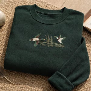 Könnte beinhalten: Dunkelgrünes Sweatshirt mit einer gestickten Darstellung von zwei Enten im Flug über Schilf. Das Sweatshirt liegt gefaltet auf einer strukturierten Oberfläche, mit einer Feder und einer Vase mit Trockenblumen in der Nähe.