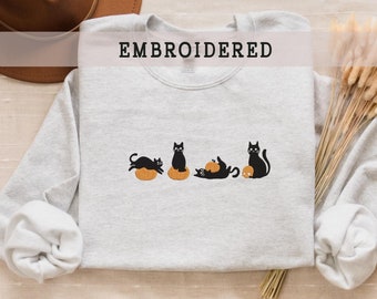 May include: A gray sweatshirt with the word "EMBROIDERED" printed on the front. The sweatshirt features an embroidered design of five black cats sitting on orange pumpkins.