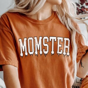 May include: A burnt orange t-shirt with the text "MOMSTER" in white dripping letters. The shirt is worn by a person sitting on a bed with a white blanket.