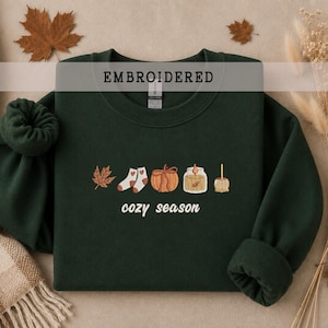 Könnte beinhalten: Ein dunkelwaldgrüner Rundhals-Pullover mit einer Stickerei, die ein Ahornblatt, ein Paar Socken, einen Kürbis, eine Kerze und einen kandierten Apfel zeigt. Unter den Symbolen steht der Text "cozy season" in einer weißen, kursiven Schrift.
