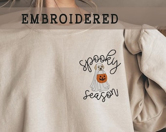 May include: A beige sweatshirt with an embroidered design of a dog dressed as a ghost holding a pumpkin. The text "Spooky Season" is embroidered below the dog.