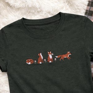 Könnte beinhalten: Dunkelgrünes T-Shirt mit einem gestickten Design von Füchsen in verschiedenen Posen und kleinen weißen Blumen. Die Füchse sind braun und weiß. Das Shirt wird auf einer weißen, flauschigen Oberfläche präsentiert.