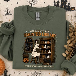 Może przedstawiać: Oliwkowa bluza z okrągłym dekoltem z nadrukiem graficznym zawierającym tekst "WELCOME TO OUR HAUNTED LIBRARY" oraz "SPOOKY STORIES THAT WON'T REST". Ilustracja przedstawia ducha czytającego książkę na drabinie obok regału z książkami.