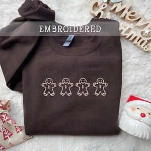 Może przedstawiać: Brązowa bluza z białym haftem przedstawiającym czterech piernikowych ludzików. Tekst "EMBROIDERED" znajduje się nad haftem.