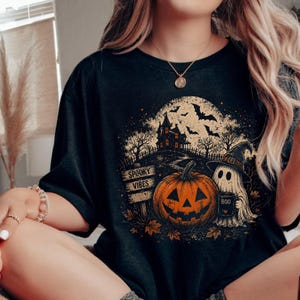 Peut inclure: Un t-shirt noir avec une impression graphique représentant une citrouille d'Halloween, un fantôme portant un chapeau de sorcière tenant une tasse marquée "BOO", une maison hantée, des chauves-souris et un panneau en bois "SPOOKY VIBES" devant une lune.