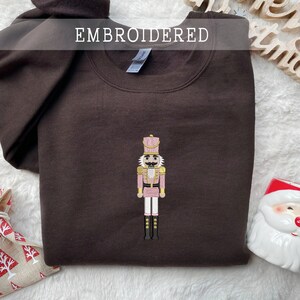Op de afbeelding: Een bruine geborduurde sweatshirt met een notenkrakerontwerp in roze, goud en zwart. Het woord "EMBROIDERED" staat bovenaan. Kerstversieringen, waaronder een Santa-mok en een ster, zijn zichtbaar.