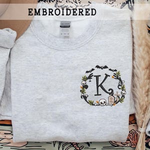 Könnte beinhalten: Hellgraues besticktes Sweatshirt mit einem Halloween-Kranz-Design. Der Kranz enthält Fledermäuse, Totenköpfe und den Buchstaben "K". Das Wort "GESTICKT" ist über dem Sweatshirt aufgedruckt.