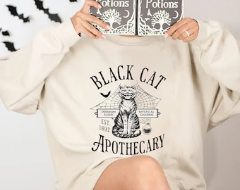 May include: A beige sweatshirt featuring a black cat graphic and the text "Black Cat Apothecary". The words "Premium Elixir" and "Mystical Charms" are on either side of the cat. The cat is sitting in a spiderweb with a bat flying above it. The text "Est. 1692" is below the cat.