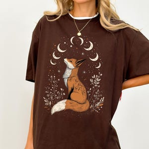 K&ouml;nnte beinhalten: Braunes T-Shirt mit einer Fuchsgrafik. Der Fuchs ist orange und wei&szlig; und blickt auf ein Mondsichel-Design. Das Shirt hat eine lockere Passform und kurze &Auml;rmel, mit einem sichtbaren wei&szlig;en Kragen. Das Design hat ein himmlisches Thema.