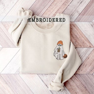 Könnte beinhalten: Cremefarbenes Sweatshirt mit einem gestickten Geisterdesign. Der Geist trägt eine orangefarbene Mütze und hält einen Kaffeebecher und einen geschnitzten Kürbis. Das Wort "EMBROIDERED" ist oben auf dem Sweatshirt aufgedruckt.