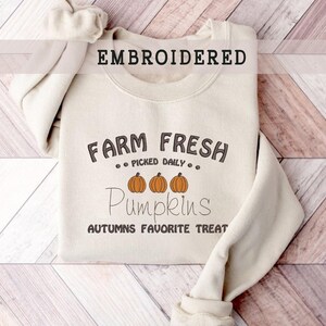 Op de afbeelding: Een cr&egrave;mekleurige sweatshirt met het woord "EMBROIDERED" bovenaan. De sweatshirt heeft de tekst "FARM FRESH PICKED DAILY" boven drie oranje pompoenillustraties en het woord "Pumpkins" in een scriptlettertype. De zin "AUTUMNS FAVORITE TREAT" staat eronder.