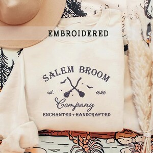 K&ouml;nnte beinhalten: Ein cremefarbener Pullover mit dem Text "Salem Broom Company", der in Schwarz gestickt ist. Der Text ist von einem Kreis mit den Worten "est. 1626" und zwei gekreuzten Besen umgeben.