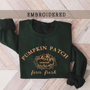 Könnte beinhalten: Ein dunkelgrüner Pullover mit dem Text "Pumpkin Patch" in goldener Stickerei. Der Text ist über einem Design aus drei Kürbissen mit Blättern und dem Wort "seasonal" im Inneren gewölbt. Unter den Kürbissen befindet sich der Text "farm fresh".