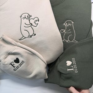May include: Two sweatshirts, one beige and one olive green, featuring embroidered otter designs. The beige sweatshirt has an otter holding a heart, while the green one has an otter with a different heart design. Sleeves are embroidered with initials and a date.
