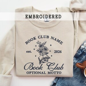 Könnte beinhalten: Ein beiges, besticktes Sweatshirt mit dem Text "EMBROIDERED" oben. Das Sweatshirt zeigt ein Buchclub-Design mit dem Text "BOOK CLUB NAME" und "Book Club OPTIONAL MOTTO" mit einer floralen Illustration. Eine Jeans und eine Tasse sind neben dem Sweatshirt.