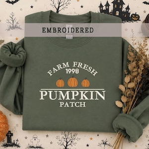 Może przedstawiać: Oliwkowozielona bluza z okrągłym dekoltem z białym haftowanym napisem FARM FRESH 1998 PUMPKIN PATCH, uzupełnionym trzema małymi pomarańczowymi ilustracjami dyń na środku klatki piersiowej. Odzież posiada długie rękawy z prążkowanymi mankietami.