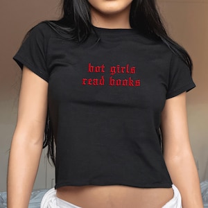 Puede incluir: Una camiseta corta negra de manga corta con el texto "hot girls read books" bordado en letras góticas rojas en el pecho. La prenda tiene un cuello redondo y un dobladillo recto que se sitúa por encima de la cintura.