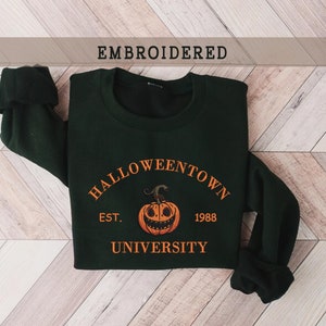 May include: A dark green sweatshirt with an embroidered design. The design features a pumpkin with a face, the text "Halloweentown University", and the years "EST. 1988".
