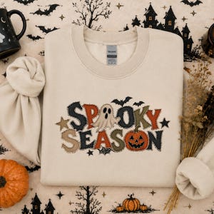 Peut inclure: Un sweat-shirt à col rond couleur crème avec un motif brodé affichant le texte "SPOOKY SEASON" dans un style patchwork. Le lettrage intègre un fantôme blanc, une citrouille d'Halloween orange ainsi que de petites étoiles et chauves-souris noires.