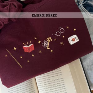Könnte beinhalten: Burgunderfarbenes Sweatshirt mit gestickten magischen Motiven. Es zeigt einen Zauberstab, ein offenes Buch, eine Brille, einen Schlüssel, einen Umschlag und Sterne. Das Wort "EMBROIDERED" steht über dem Design.