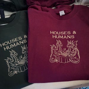 Könnte beinhalten: Zwei T-Shirts, eines in Dunkelgrün und eines in Burgunderrot, mit dem Text "HOUSES & HUMANS" und einem weißen Stickdesign von Drachen, die ein Brettspiel spielen. Die T-Shirts sind gefaltet.