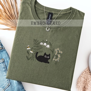Może przedstawiać: Oliwkowo-zielona koszulka z haftowanym wzorem przedstawiającym czarnego kota, kwiaty i pszczołę. U góry wydrukowano napis "EMBROIDERED". Widoczny jest również srebrny naszyjnik z zawieszką w kształcie słońca.