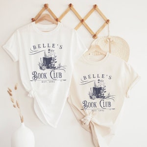May include: Two white t-shirts with a navy blue graphic design. The graphic features a book with flowers and the text "Belle's Book Club Est. 1991".