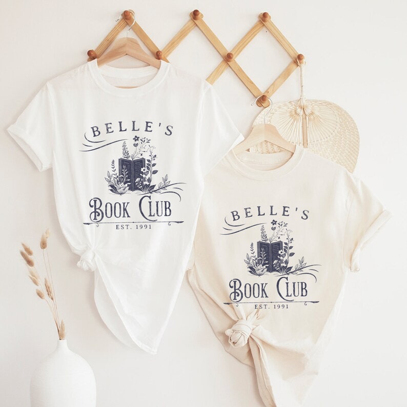 May include: Two white t-shirts with a navy blue graphic design. The graphic features a book with flowers and the text "Belle's Book Club Est. 1991".