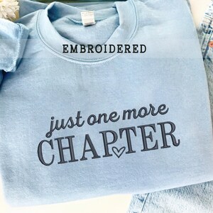 Pode incluir: Camisola azul clara bordada com as palavras "just one more CHAPTER" em escrita cinza escuro. A palavra "EMBROIDERED" est&aacute; acima da gola. Um pequeno cora&ccedil;&atilde;o est&aacute; abaixo da palavra "CHAPTER".