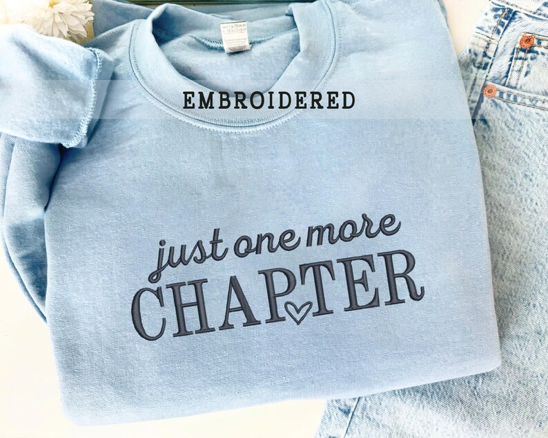 Pode incluir: Camisola azul clara bordada com as palavras "just one more CHAPTER" em escrita cinza escuro. A palavra "EMBROIDERED" est&aacute; acima da gola. Um pequeno cora&ccedil;&atilde;o est&aacute; abaixo da palavra "CHAPTER".