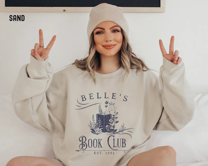 May include: A beige sweatshirt with a graphic design of an open book with flowers and the text "Belle's Book Club Est. 1991".