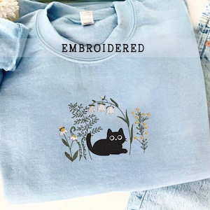 Può includere: Una felpa azzurra con la scritta "EMBROIDERED" in alto. La felpa presenta un disegno ricamato di un gatto nero sdraiato tra fiori e verde. Il gatto ha grandi occhi rotondi.