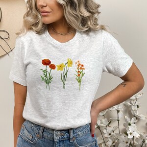 May include: A gray t-shirt with a floral print featuring orange, yellow, and white flowers. The flowers are arranged in a row across the chest of the shirt.