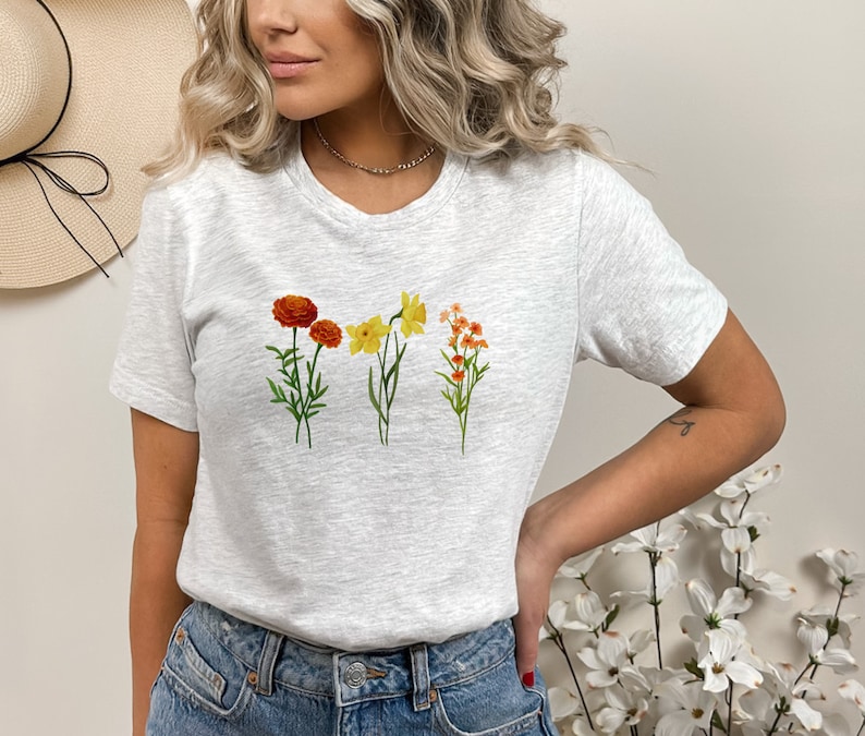 May include: A gray t-shirt with a floral print featuring orange, yellow, and white flowers. The flowers are arranged in a row across the chest of the shirt.