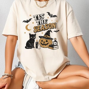 May include: Cream-colored t-shirt with a Halloween-themed graphic. The design includes a black cat, a jack-o'-lantern wearing a witch hat, bats, a crescent moon, stars, and the text "Tis the Season".