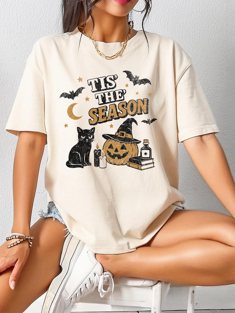 May include: Cream-colored t-shirt with a Halloween-themed graphic. The design includes a black cat, a jack-o'-lantern wearing a witch hat, bats, a crescent moon, stars, and the text "Tis the Season".