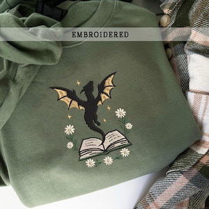 Könnte beinhalten: Olivgrünes Sweatshirt mit einem gestickten Design, das einen schwarzen Drachen mit goldenen Flügeln, ein Buch und weiße Blumen zeigt. Das Wort "EMBROIDERED" ist über dem Design aufgedruckt. Im Hintergrund ist ein kariertes Hemd zu sehen.