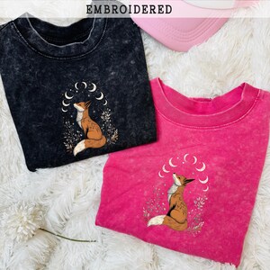 May include: Two t-shirts, one black and one hot pink, with embroidered fox designs. The fox is brown and white, surrounded by crescent moons and foliage. A pink and white trucker hat is also visible. The word "EMBROIDERED" is displayed.