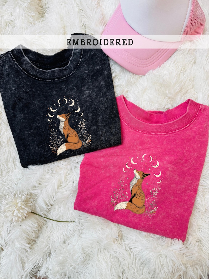 May include: Two t-shirts, one black and one hot pink, with embroidered fox designs. The fox is brown and white, surrounded by crescent moons and foliage. A pink and white trucker hat is also visible. The word "EMBROIDERED" is displayed.